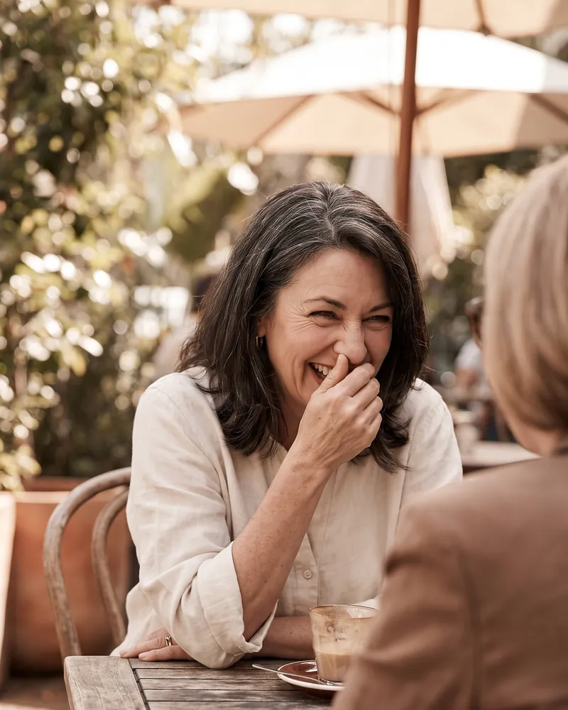 A woman at a cafe table laughing with a friend and covering her mouth with her hand